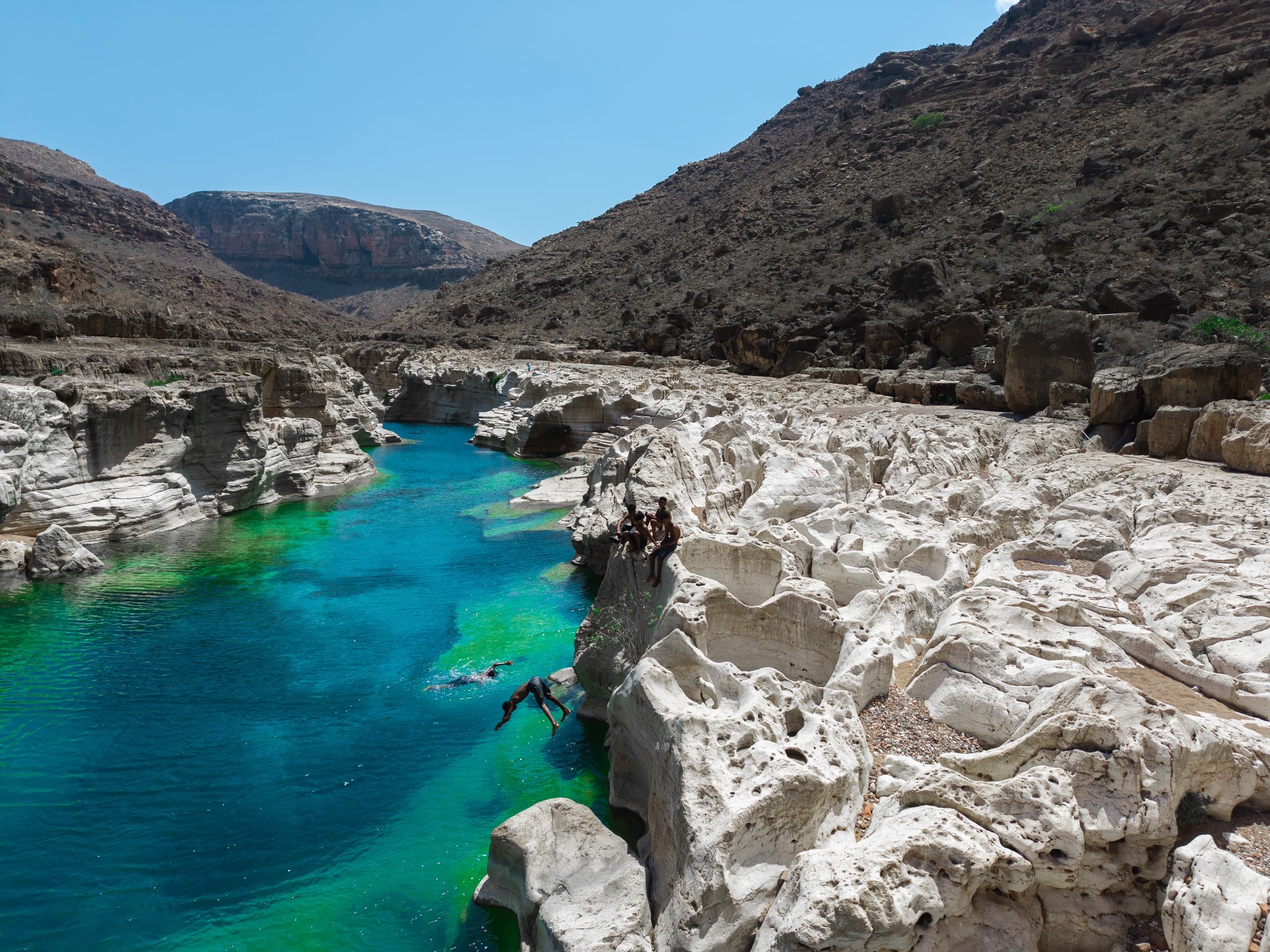 Local boys leaping into an emerald freshwater pool in a white limestone wadi