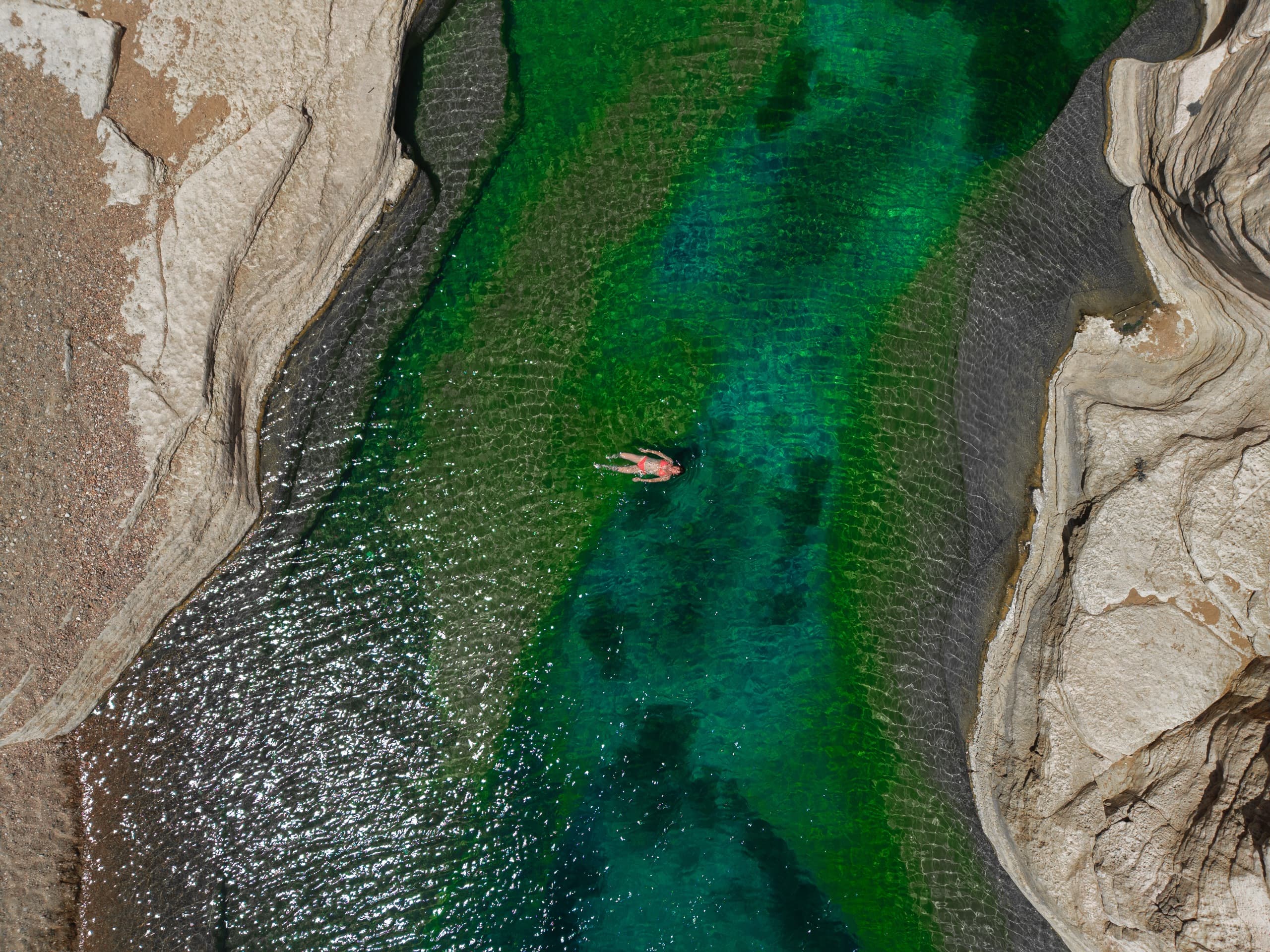 A person floating in a clear emerald wadi pool, seen from directly above